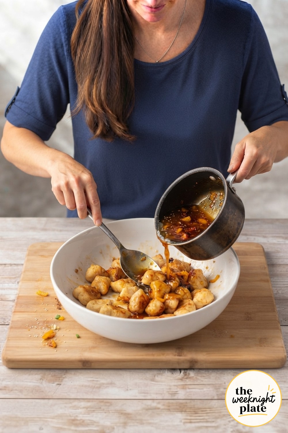 Glossy orange sauce being poured over crispy baked chicken pieces in a mixing bowl.